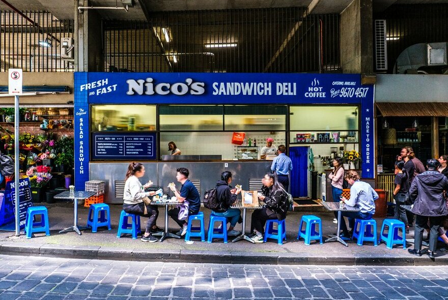 People eating on blue stools outside a blue hole in the wall sandwich shop.