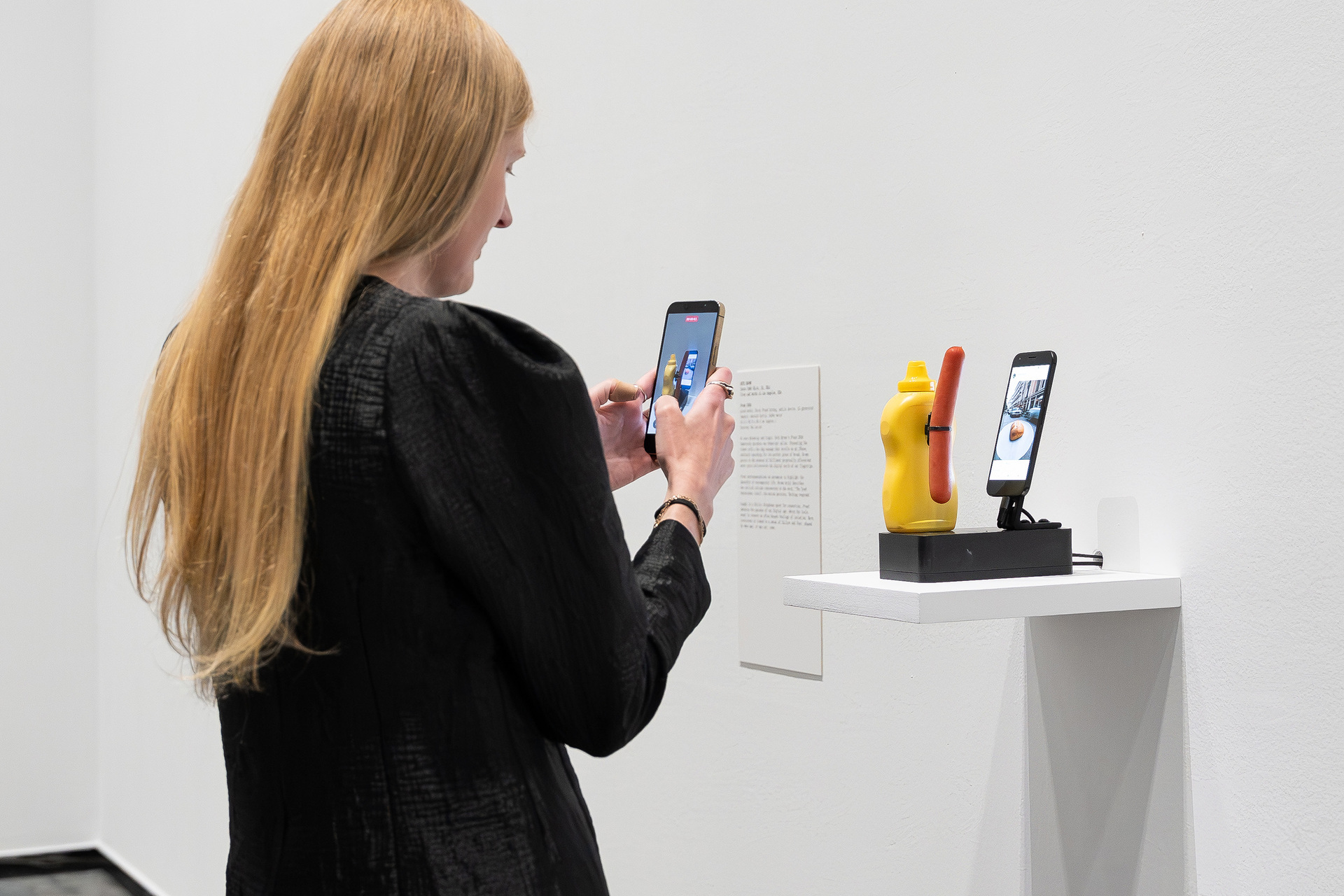 A person is taking a photo of an art installation featuring a bottle of mustard holding a sausage scrolling on a phone