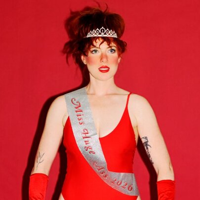 Australian comedian and actor Frankie McNair, wearing a red leotard, red gloves, a tiara and with a white sash across her torso, her hair is in a beehive style and she stands in front of a red background.