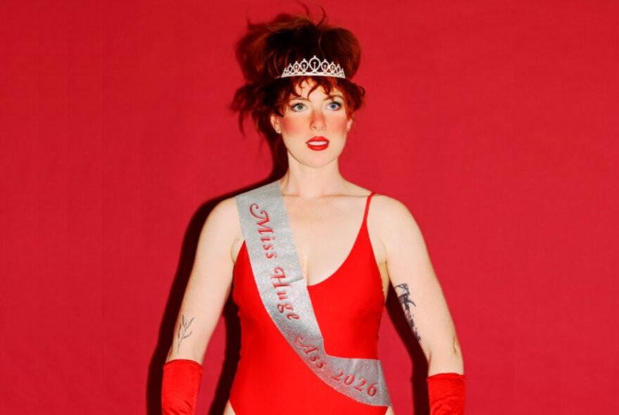 Australian comedian and actor Frankie McNair, wearing a red leotard, red gloves, a tiara and with a white sash across her torso, her hair is in a beehive style and she stands in front of a red background.