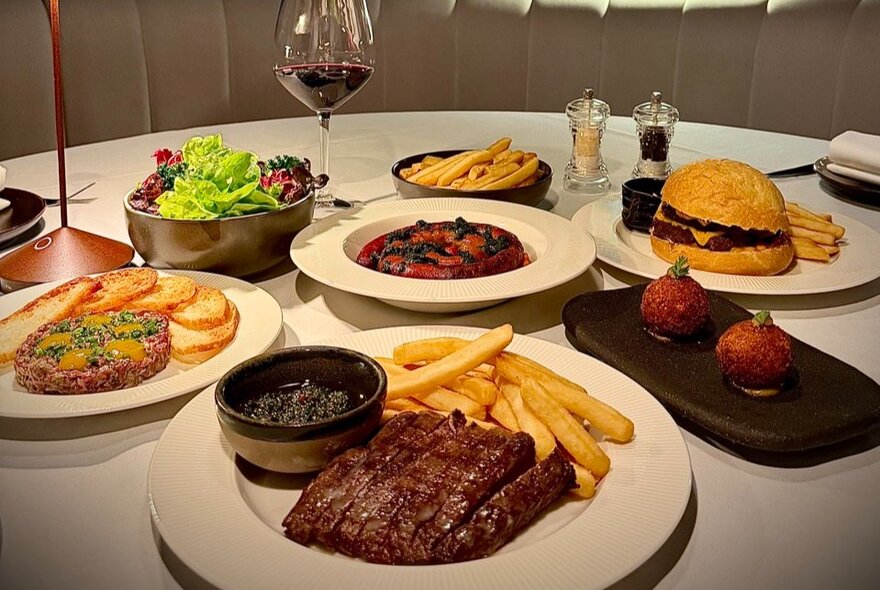 A large spread of modern steakhouse dishes, including a chargrilled steak with fries and sauce, a gourmet burger, steak tartare, salad, and small bites.