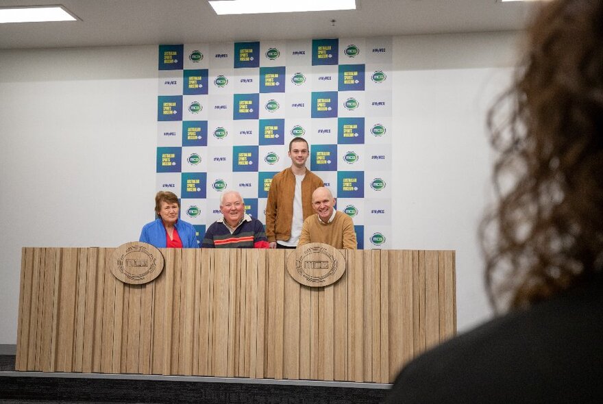 Four people sitting at a wooden desk with a person's hair visible in the foreground. 