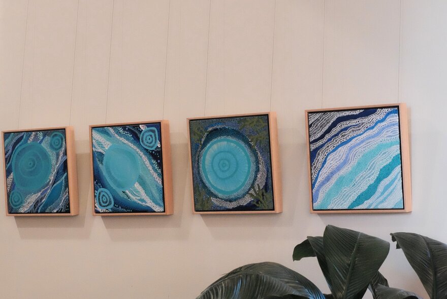 Four square, contemporary Indigenous Australian paintings featuring shades of blue and teal hang in a row on a plain, neutral wall.