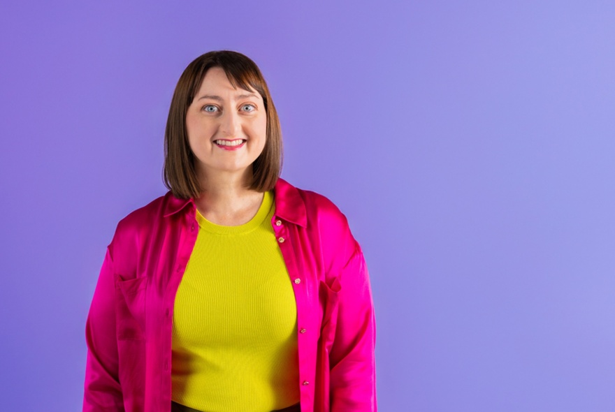 Comedian Sonia Di Iorio wearing yellow and pink, against a purple background.