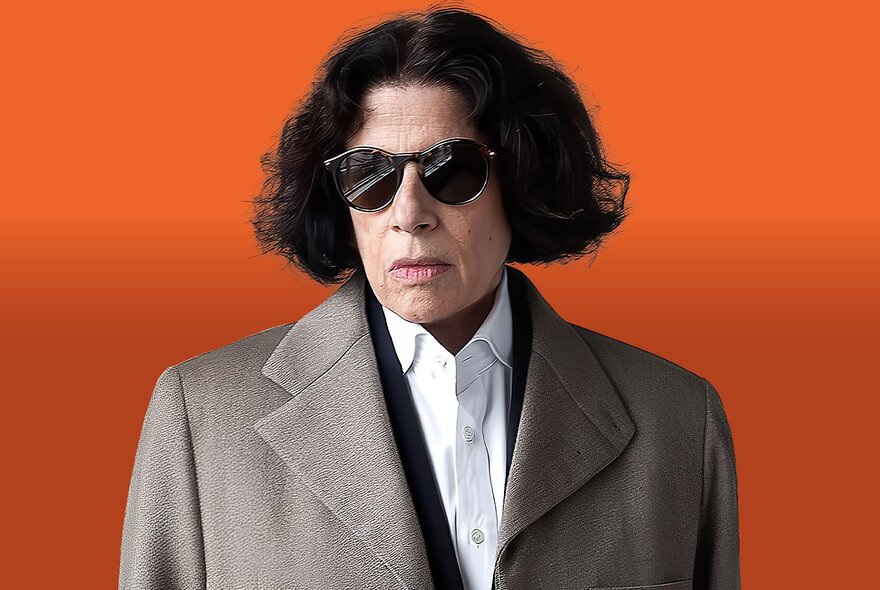 Fran Lebowitz wearing a white shirt, overcoat, and dark sunglasses with a neutral expression, posed against an orange background.