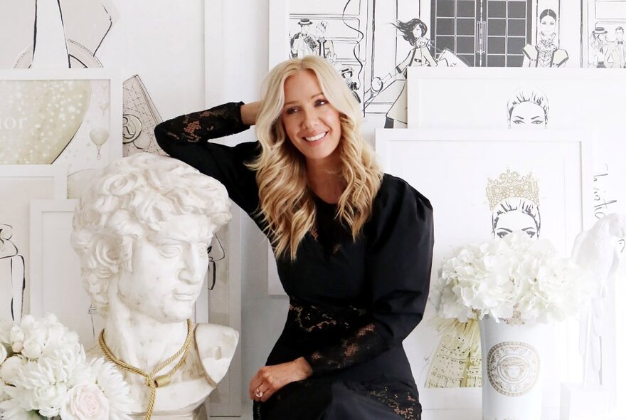 Author and illustrator Megan Hess, smiling and wearing a black dress, long blond hair, and seated in a studio of her illustrations and surrounded by white props including a white ceramic bust, a vase of white flowers and other objects.