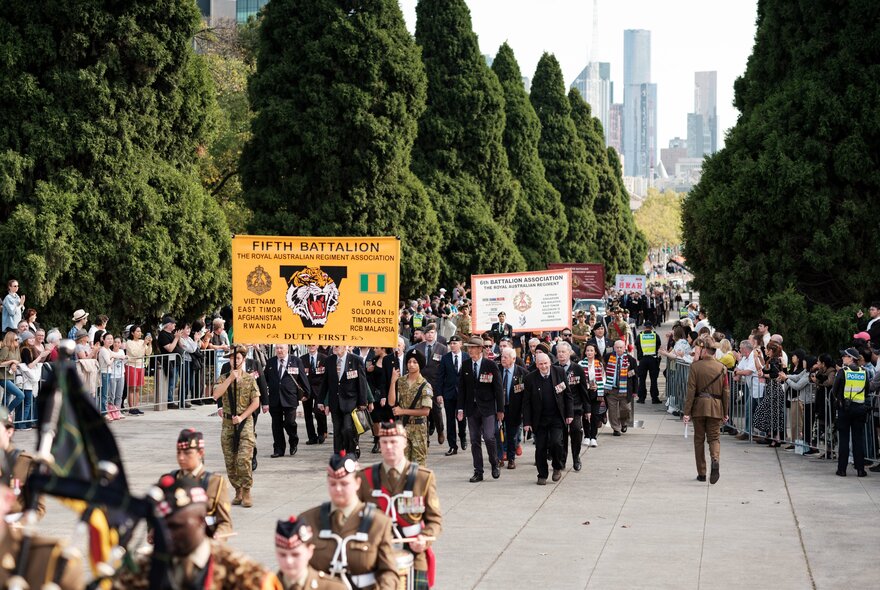 A parade of returned servicemen and women marching in front of a crowd, holding banners.