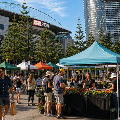 An outdoor farmers' market located in the Docklands area, with stalls selling fresh produce, people wandering around, and Marvel Stadium in the background.