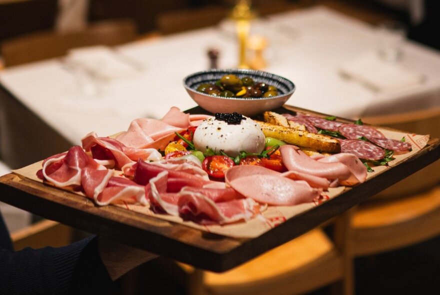 An antipasto wooden platter of sliced hams and salami, cheese and olives.