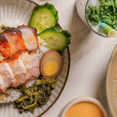 Two plates of Indonesian food including pork belly, rice, cucumber, egg, water spinach and noodles, and a glass of es cendol (iced sweet dessert).