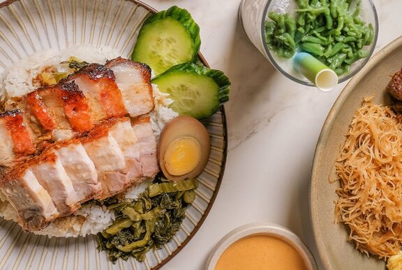 Two plates of Indonesian food including pork belly, rice, cucumber, egg, water spinach and noodles, and a glass of es cendol (iced sweet dessert).