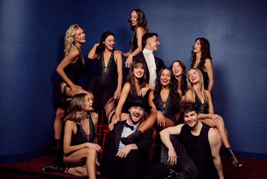A group shot of the dancers and performers at Viva club, smiling and looking at each other, some seated on the floor, others standing, posed against a blue backdrop.