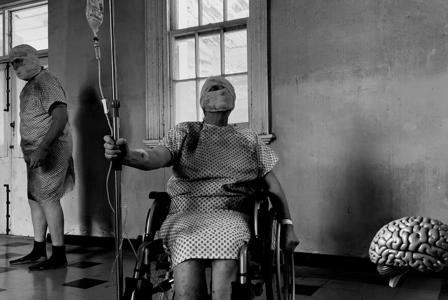 Black and white still from the movie Cerebral Organoid depicting a surreal or staged hospital scene with figures wrapped in hospital gowns, their faces covered in bandages, one in a wheelchair attached to a drip and another walking around in the space.