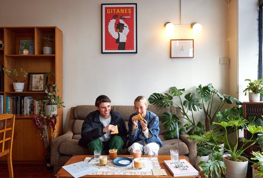 A couple sitting on a couch in a leafy cafe enjoying coffee and toasties.