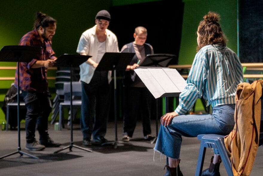 A play reading with three people standing reading before a seated person, also with a script. 