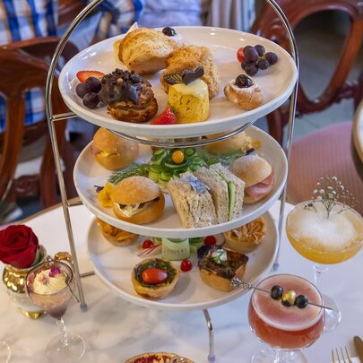 A three tiered tray of cakes and sandwiches surrounded by cocktails.