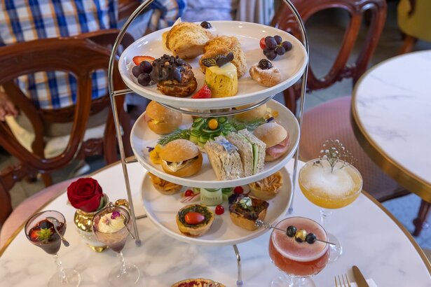 A three tiered tray of cakes and sandwiches surrounded by cocktails.