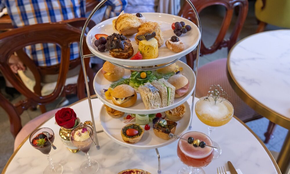 A three tiered tray of cakes and sandwiches surrounded by cocktails.