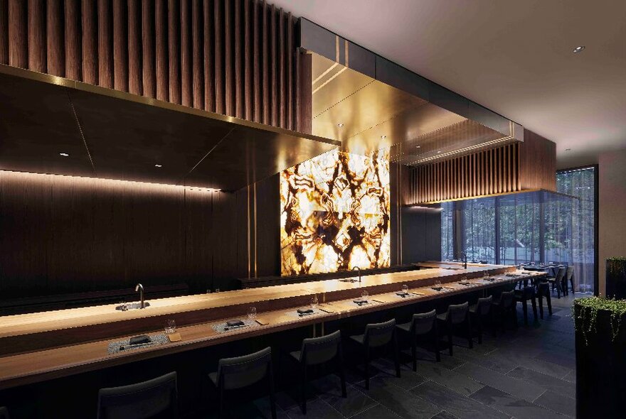 Interior of restaurant showing long dining bar area, high stools, soft down lights, an illuminated artwork, and windows to an outdoor area.