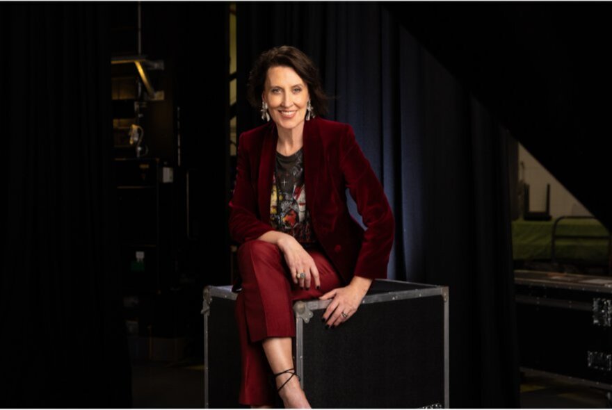 MC Virginia Trioli wearing a burgundy velvet suit jacket and dark red pants, sitting cross-legged in a studio.