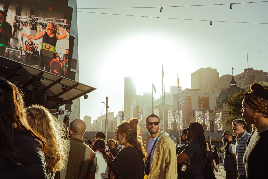 People enjoying outdoor live entertainment at Fed Square in the early evening, with city buildings in the background.