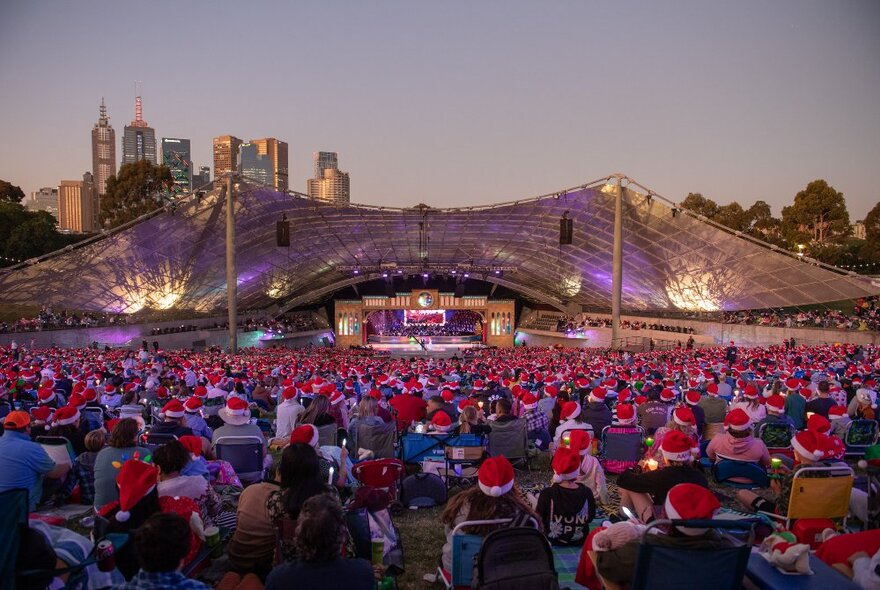 Seated crowds at Sidney Myer Music Bowl wearing Santa hats at dusk, city buildings in the distances.