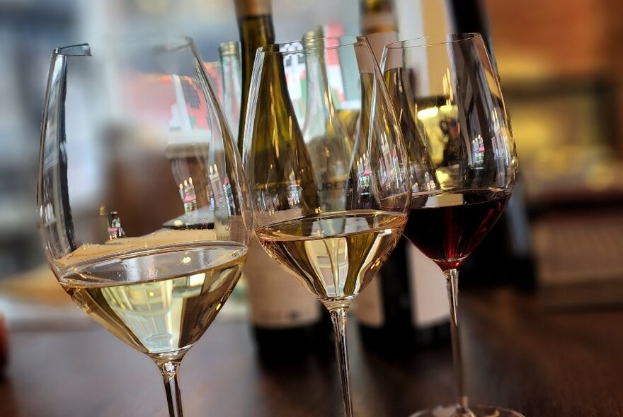 Three glasses of wine lined up on a wooden bar with half full wine bottles in the background.