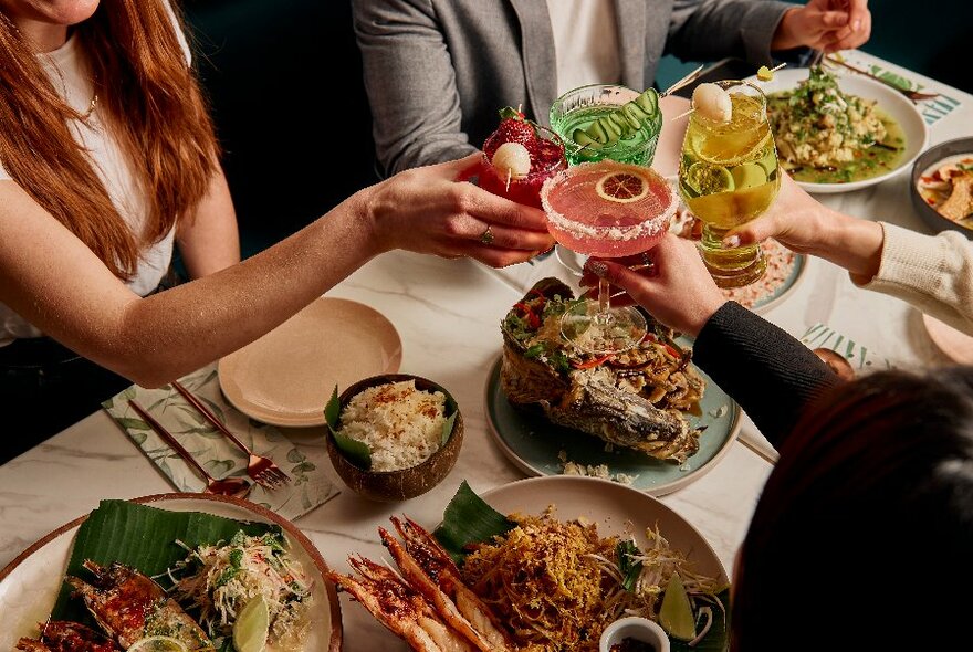 Looking down at a table filled with food dishes and people clinking cocktail glasses.