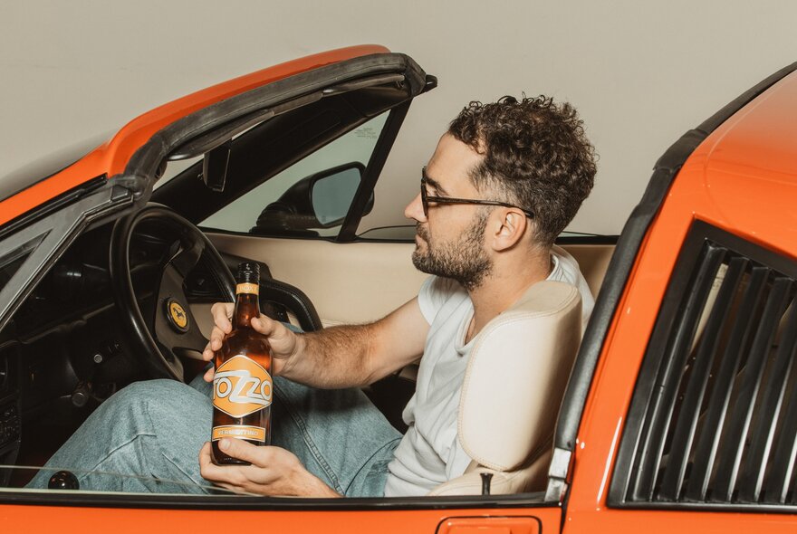 Man with beard and glasses sitting in an orange sports car holding a bottle of wine.