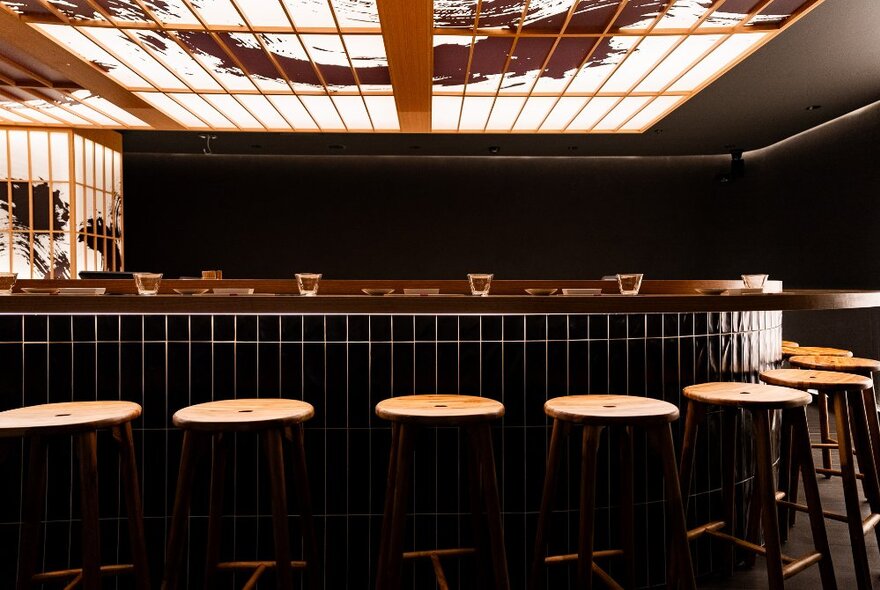 Stools lined up at a curved sushi counter topped with glasses and plates, under a white glass decorative Japanese-style ceiling.