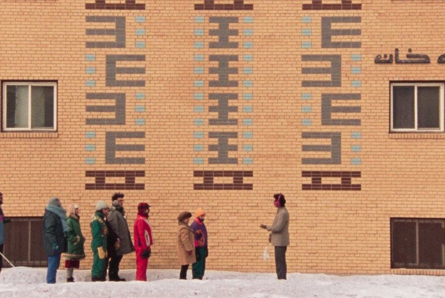 A queue of people lined up in front of a tall brick wall with coloured patterns in the brick.