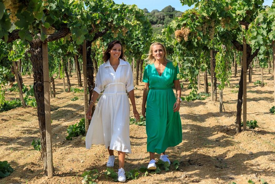 Two women walking on sandy soil in between two rows of vine plantings in a vineyard.