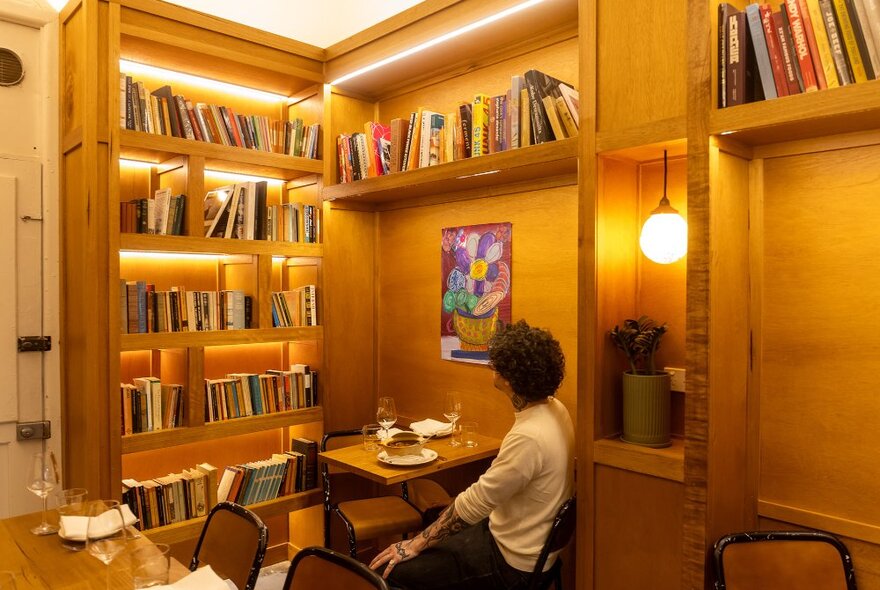 A man sitting in the corner of a wine bar looking at a bookshelf.