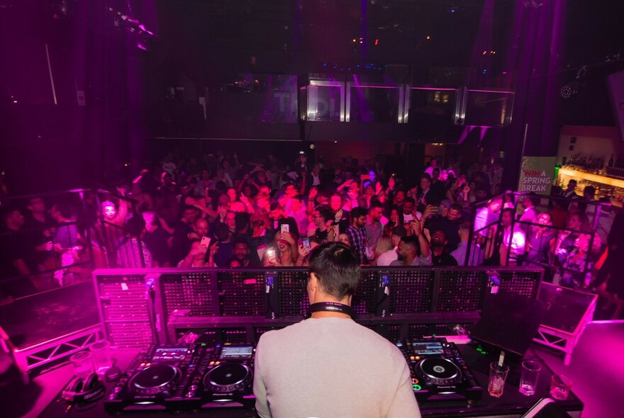 Nightclub interior with a rear view of DJ at booth producing music while a crowd in front dances.