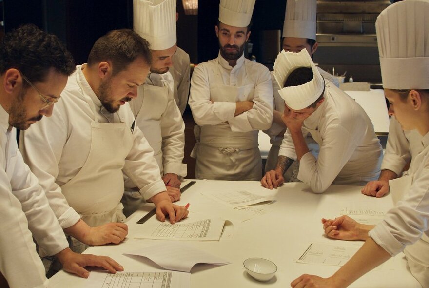 A still from the film Menus-Plaisirs – Les Troisgros that shows six chefs all wearing white chef uniforms and leaning around a table reading notes in a commercial kitchen.