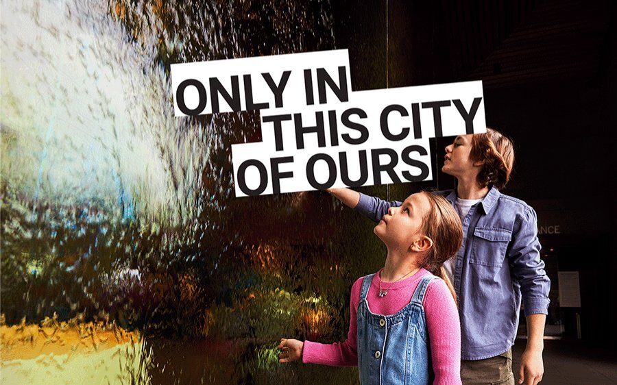 Two children looking at a glass wall water feature. Text overlay reads 'only in this city of ours'.