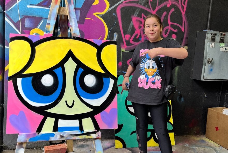 A teenager standing next to a brightly coloured canvas painting on an easel in a studio workspace. 