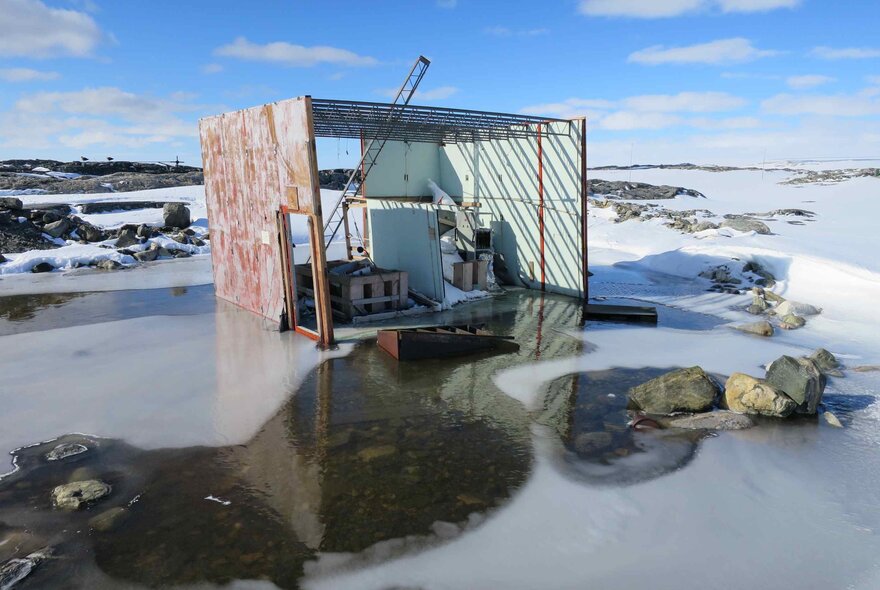 A dilapidated, rusted metal hut, possibly an abandoned research station, sitting on the edge of a frozen body of water in Antarctica, with some open dark water reflecting the structure.