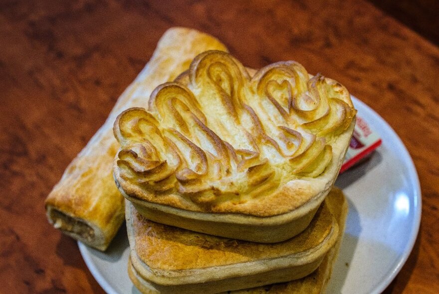 A sausage roll and two pies on a white plate on a wooden counter.