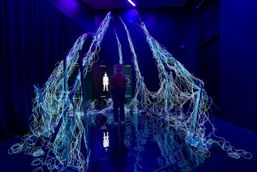 A person in a large darkened exhibition space, engaging with an immersive style installation that surrounds them, while looking at a video screen.