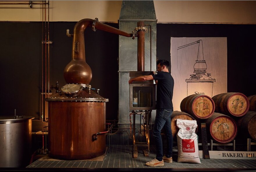 A man working a brass whisky still with whisky barrels stacked beside it.