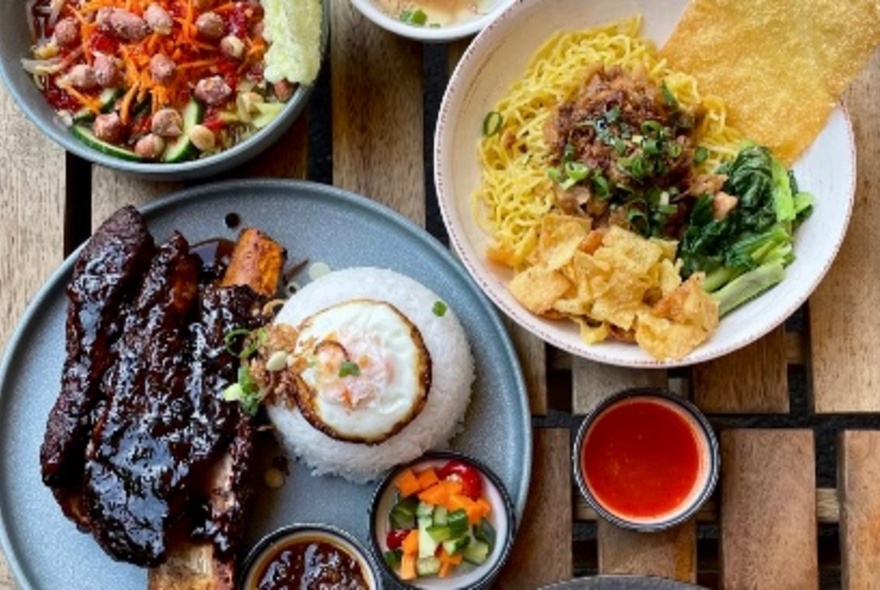 Looking down at a table filled with plates of food including large beef ribs, rice and sticky sauce, a noodle and vegetable bowl and a salad with peanuts.