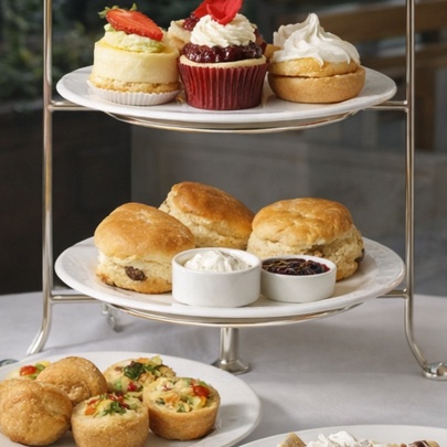 A table set for High Tea with a multi-tiered cake stand containing scones and cakes, a small plate of savoury pastries and two flutes of pink champagne.