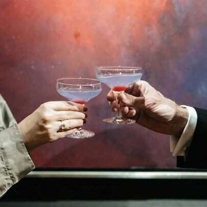 Hands opposite each other bringing two tulip cocktail glasses together in a celebratory clinking motion.