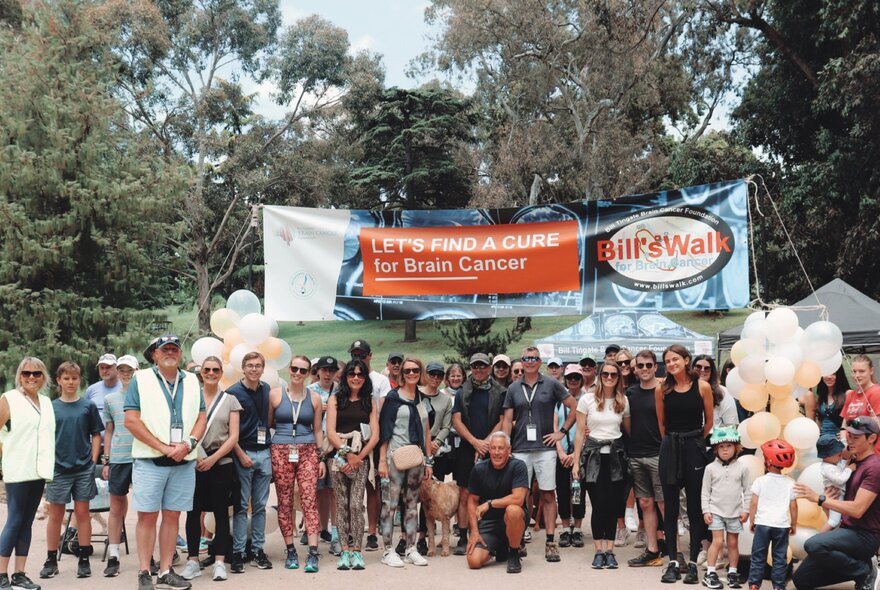 A large group of people standing outside in Kings Domain gardens, in front of a sign indicating the starting position of a charity walk.