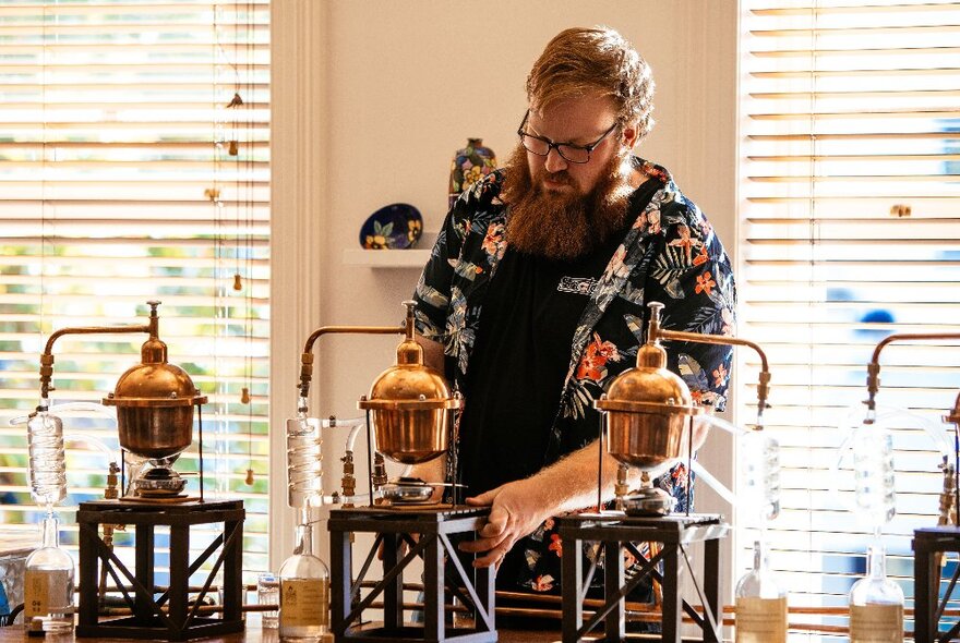 A man in a floral shirt stands behind three small copper pot gin stills on wooden frames.