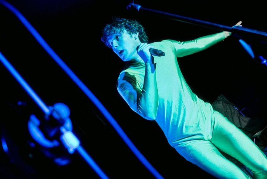 A man dressed in a green body suit, holding a microphone and performing on stage.