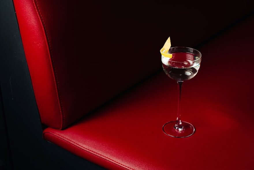 A single tall-stemmed cocktail glass with a clear cocktail and lemon twist on a red leather bench seat.