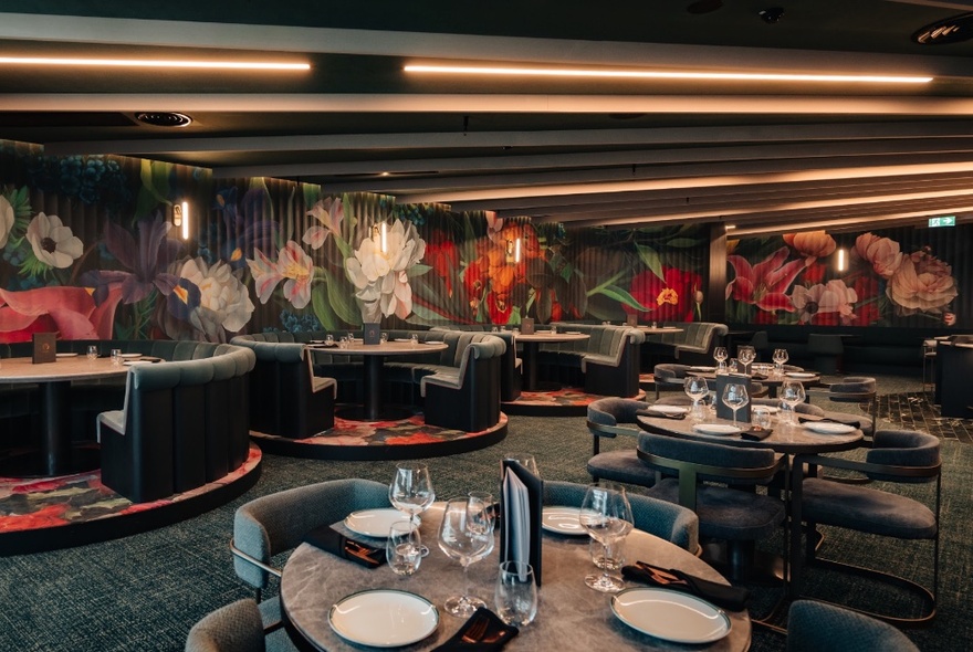 Interior of Amphora restaurant with round dining tables and chairs, in a carpeted room with flowers painted on the walls.