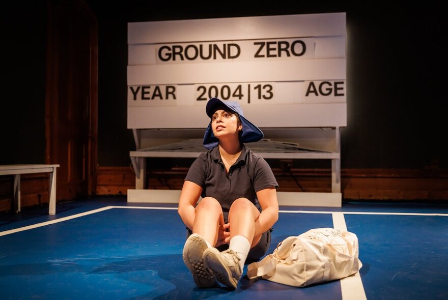 A theatre set with a child seated on a tennis court, court signage in the background.